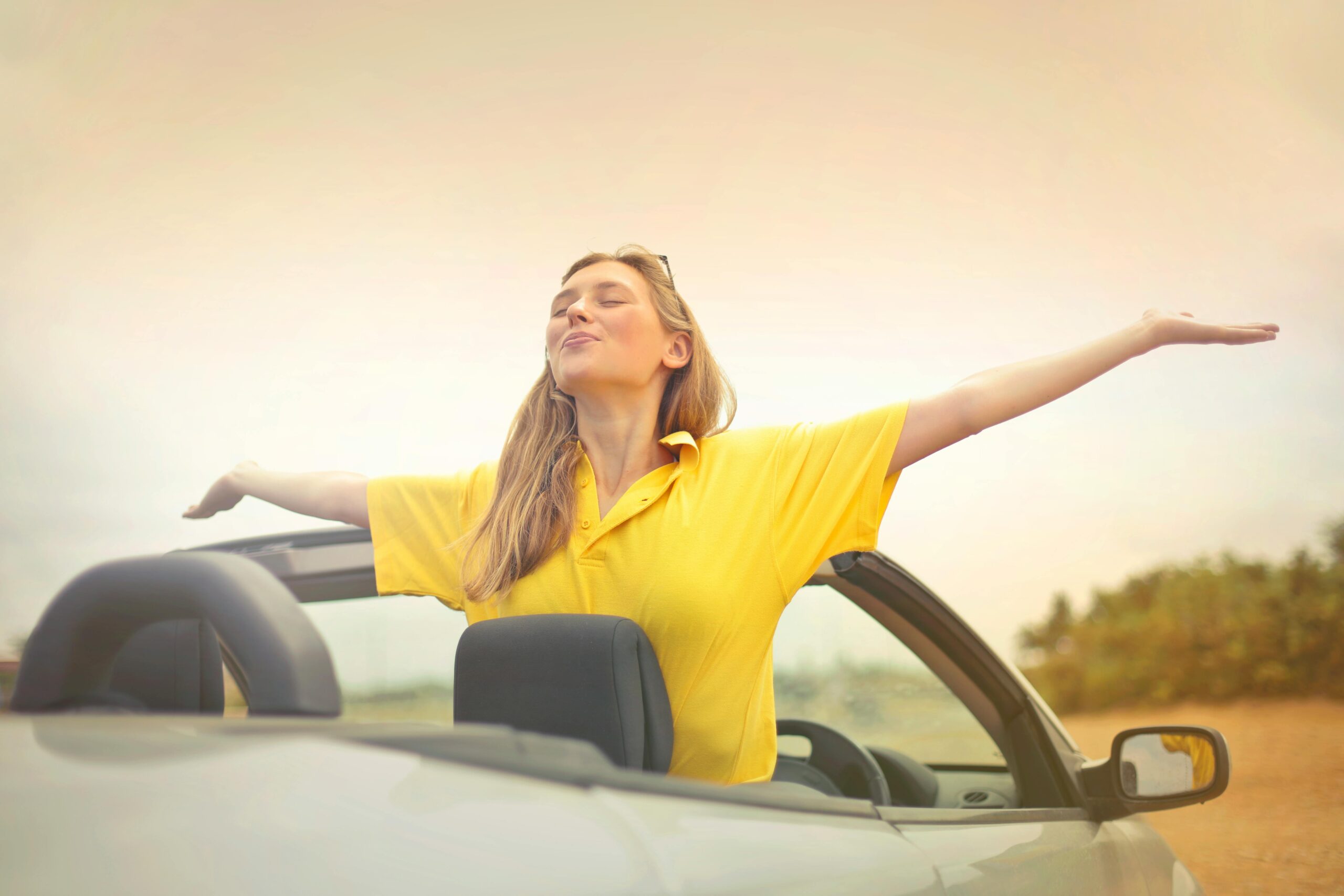 A cheerful woman in a yellow shirt enjoys freedom on a sunny drive in a convertible.